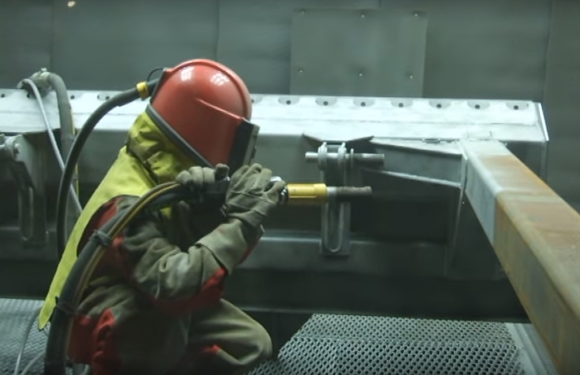 Работник завода обрабатывает дробеструем металлоконструкцию.